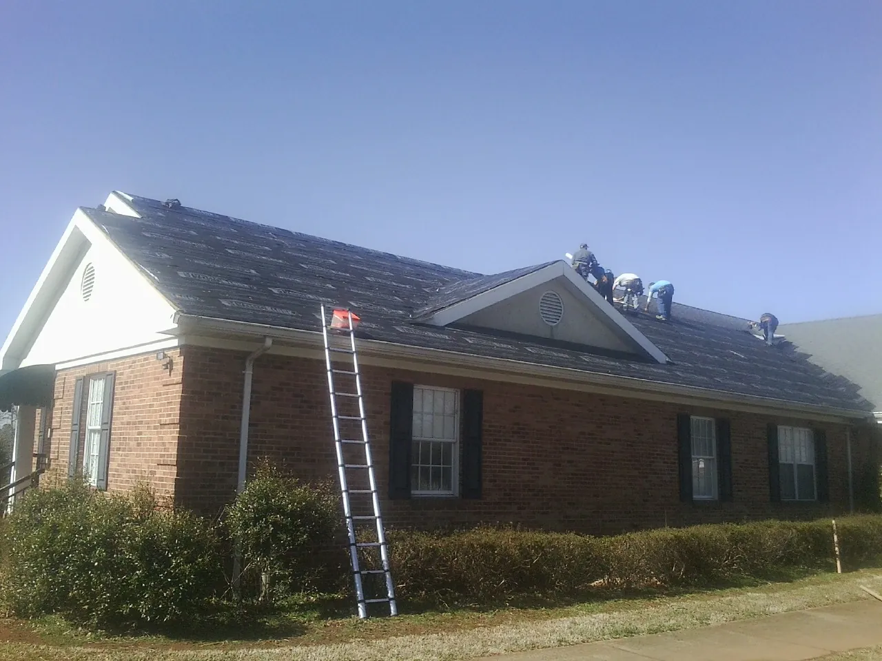 Crew tearing shingles off a brick ranch roof with a ladder set — Emergency Roof Repair work by our crew in Ashby