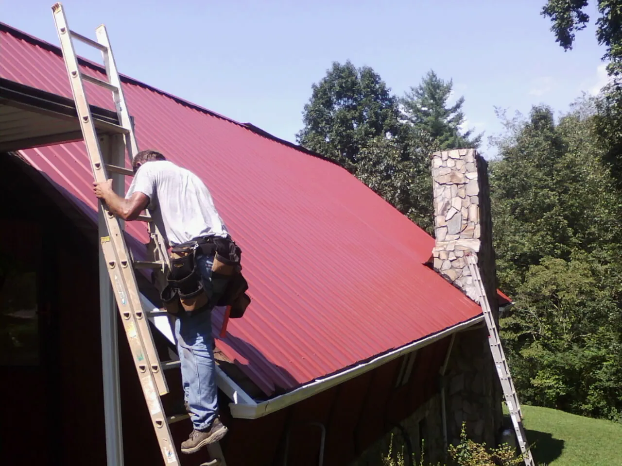 Roofer on a ladder beside a red metal roof and stone chimney — Soffit & Fascia Repair by Roofing Brothers in Ashby